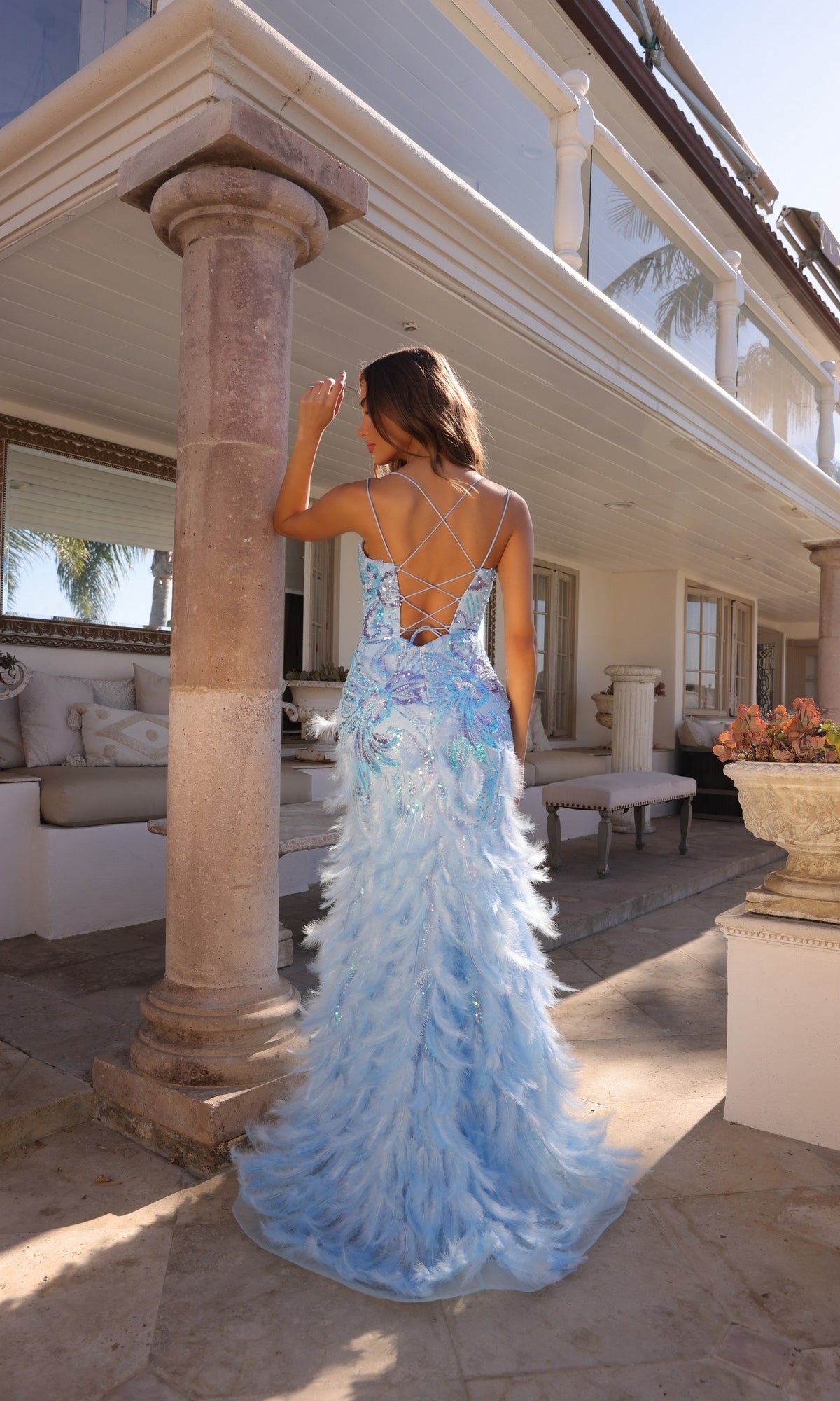 A woman wearing the Nox Anabel Feather and Sequin Prom Dress C1413 with a strappy back stands on a patio, facing away from the camera toward a house with columns and outdoor seating.